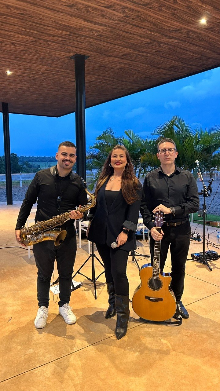 Bruna Pires entre o saxofonista e o violonista, em um espaço aberto para eventos